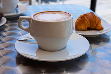 A white ceramic cup of fresh cappuccino served with a flaky sfogliatella pastry on a metallic table...