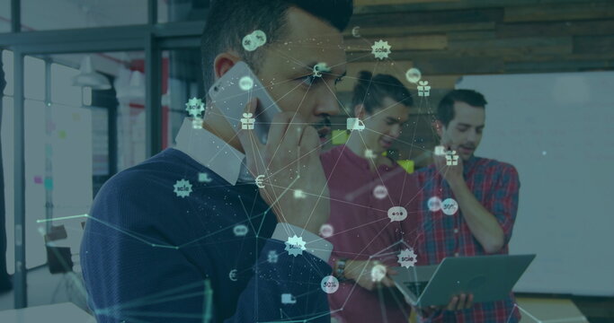 Talking businessman holding smartphone at office with coworker using laptop showing digital graphic