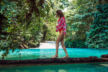 Fototapeta premium Attractive woman in bikini with a plaid shirt on a log over a turquoise river in the jungle with a waterfall. Tropical holiday adventure.