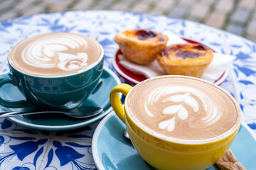 Two beautifully crafted cappuccino cups on a decorative table, accompanied by delicious pastel de nata pastries, creating a cozy coffee time atmosphere with inviting textures