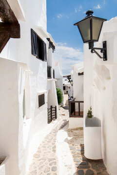 Charming narrow cobblestone street with traditional whitewashed houses and dark wooden shutters in the fishing village of Binibeca Vell, Menorca, Spain, bathed in sunlight