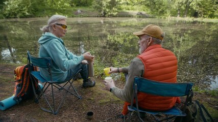 Slowmo of senior Caucasian couple of tourists sitting on camping chairs by pond and resting after long hike in forest, pouring tea from thermos bottle and enjoying scenery together - Powered by Adobe