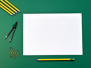 A blank sheet of paper is surrounded by yellow pencils, a compass, and paper clips on a green...