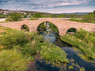 Ancient stone bridge stands majestically, arching over a river winding through a landscape of...