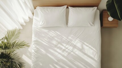 Overhead view of a neatly made bed with white linens, sunlight streaming through sheer curtains, and a bedside table with a small lamp and plant.