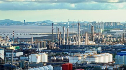 Aerial shot of coastal industrial area with an oil refinery, storage tanks, and wind turbines by the sea.