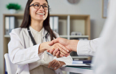 Smiling female doctor in white coat shaking hands with patient in medical office. Professional...