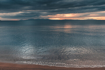 Calm sea waves gently washing sandy beach at dramatic sunset with dark clouds and islands on background