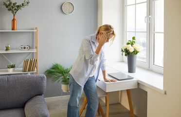 Weary blonde woman standing by window, resting hand on forehead, feeling unwell, suffering from...