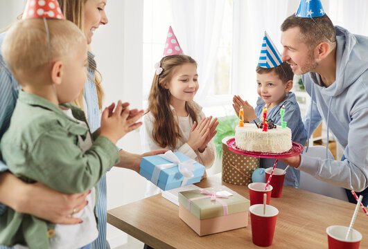 Happy smiling child girl in party hat celebrating birthday with mother, father and brothers at home, looking at cake with candles making a wish. Happy family and holiday celebration concept.