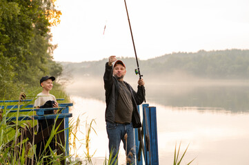 Father helping his son prepare fishing gear on a misty morning by the lake, showing patience and...