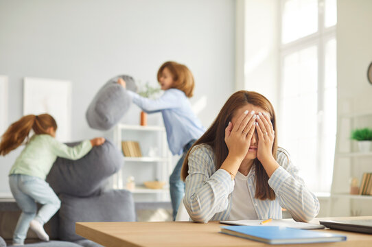 Stressed tired mother sitting at desk with her face in her hands, trying to concentrate on work while her two noisy playful children shouting, jumping and having pillow fight on sofa in background.