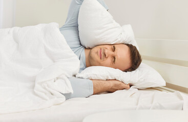 Stressed young man lying in bed, pressing pillow over his head with pained expression on his face....