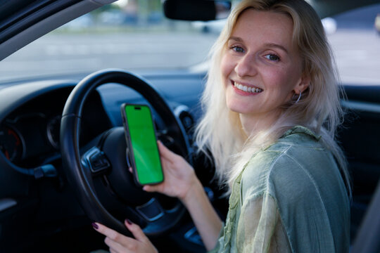 A Young Woman in a Car Holding a Smartphone with a Green Screen, Smiling Cheerfully, Capturing the Joy of Modern Technology and Life