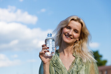 Smiling Woman Holding a Water Bottle Against a Bright Blue Sky with Fluffy Clouds, Emphasizing the...