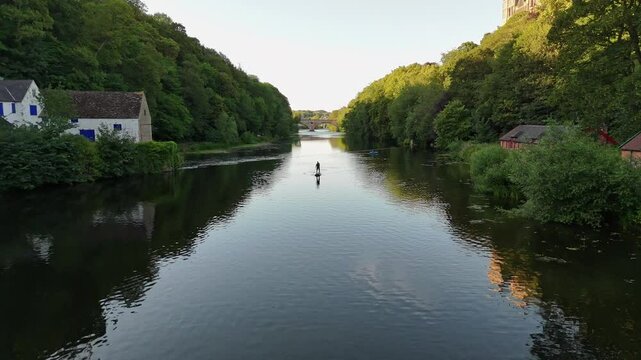 A person stand up padle boarding in the river in Durham, United Kingdom. Sunset and trees around the water. Aerial View