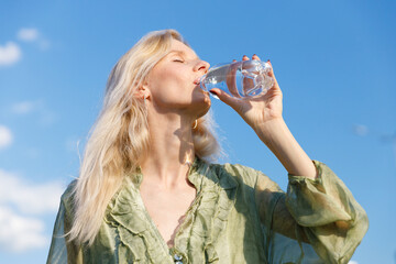 Refreshing Hydration: Woman Enjoys a Clear Glass of Water Beneath a Bright Blue Sky on a Sunny Day,...