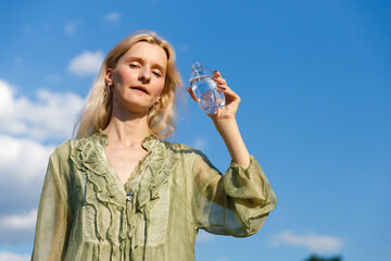 A Young Woman Holding a Clear Water Bottle Against a Stunning Blue Sky with Soft Clouds in the...