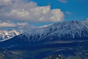 Snowy mountain range under partly cloudy sky; vibrant forested foreground adds depth and contrast to the serene alpine scene.