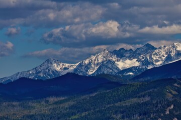 Snowy peaks rise beneath overcast sky; forested hills and rugged slopes frame a serene, windswept...