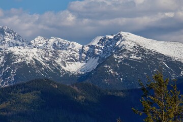 Snow-covered peaks rise above forested ridges beneath scattered clouds; natural layers evoke depth, serenity, and crisp alpine air.