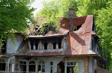 Neglected house with rusted roof and broken windows stands overgrown in lush greenery; age and...