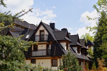 Three-gabled wooden home with light walls and dark roof sits in lush greenery; multiple balconies offer elegant rustic charm