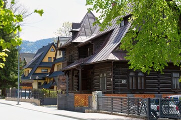 Dark brown house with metal roof and fenced yard sits in wooded neighborhood; green trees and...