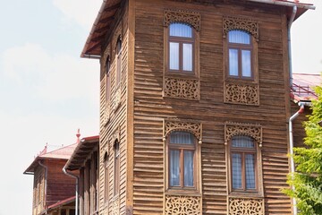 Ornately carved wooden house with arched windows and rustic charm; second similar building peeks in, framed by trees and sky