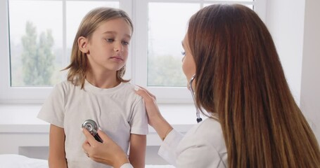 Portrait of smiling sick child patient sitting in clinic for a medical checkup with friendly doctor. Female pediatrician with stethoscope examining kid's heartbeat at hospital. 4k video. - Powered by Adobe