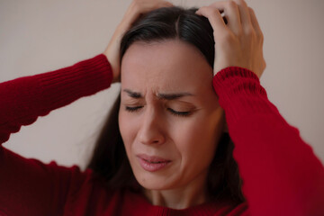 Young woman in a red sweater holding her head with both hands, eyes closed, expressing stress,...