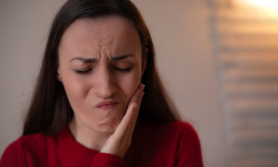 Young woman in a red sweater touching her cheek with a pained expression, suffering from toothache...