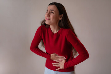 Young woman in a red sweater holding her stomach with a pained expression, suffering from stomach ache or abdominal pain while standing indoors.