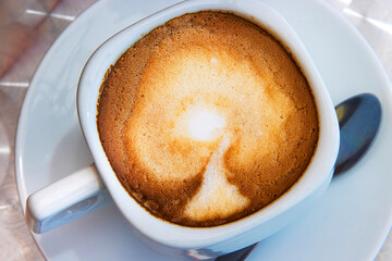 Closeup view of a cappuccino with creamy milk foam in a square white ceramic cup, placed on a...