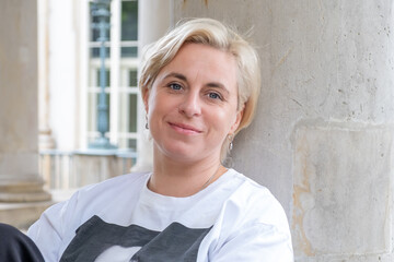 Midlife woman with short blonde hair, wearing a casual white shirt, smiles warmly while leaning...