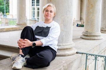 Midlife woman with short blonde hair sits comfortably on stone steps, wearing casual attire,...