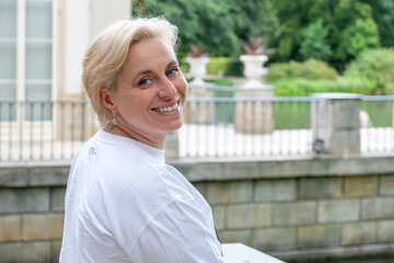 Midlife woman with short blonde hair smiles warmly while enjoying a serene outdoor setting,...