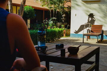 A person relaxes at an outdoor seating area under sunlight, while a shadow is cast on a wall depicting a seated cowboy wearing a hat, in a tranquil and inviting tropical environment.