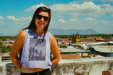 Happy young woman with sunglasses posing outdoors on a sunny rooftop with a scenic background of a historic town and mountains. Taken on 35mm film in León, Nicaragua.