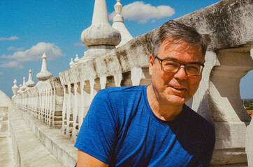 A man wearing glasses smiles to the camera while sitting on a white historic building rooftop, surrounded by architectural beauty and basking under a bright blue sky. Taken on 35mm film.