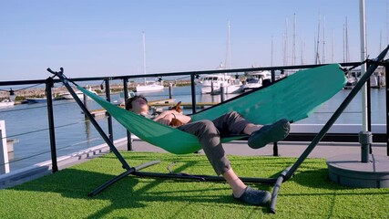 Teenager relaxing in teal hammock with small dog, overlooking marina serene waterfront on bright sunny day, embodying peaceful outdoor lifestyle and leisure moment