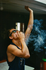 A stylish man smoking a cigar indoors while smoke swirls around him, creating a dramatic and moody setting enhanced by contrasting lighting and composition. Taken on 35mm Film.