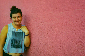 Stylish young woman smiling comfortably next to a brightly colored pink wall, wearing a casual outfit featuring a tank top printed with tropical motifs. Taken on 35mm Film.