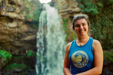 A cheerful woman wearing a blue shirt stands before a stunning waterfall surrounded by lush greenery, portraying joy and adventure in an outdoor natural environment. Taken on 35mm Film.