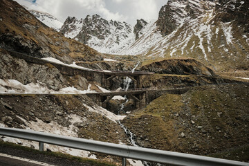 Stunning mountain landscape with a winding road and double bridge. Snow-capped peaks and a cascading waterfall create a dramatic scene. Perfect for travel, nature, or adventure themes.