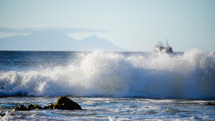 Dramatic ocean wave crashing near shore with splashes in focus and fishing boat blurred on the...