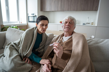 Struggling upset aged man holding thermometer wrapped in shawl check fever with concerned wife on...