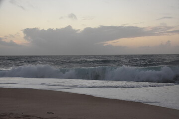 Waves crash onto the shore at Praia do Norte in Nazaré, Portugal, under a cloudy sunset...