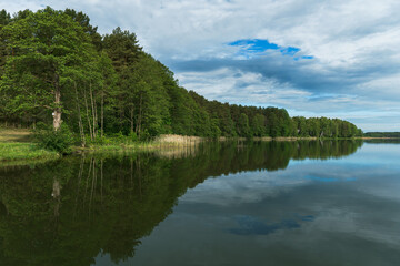 Sunny summer landscape on the lake. Reflection of the sky and clouds in the water. A valley of green trees on the shore of a lake. A concept for travel and camping.