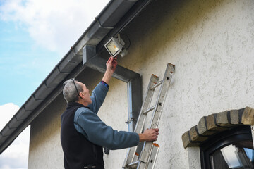 Electrician reaches for motion sensor LED floodlight while standing on ladder installing it on facade of house under gutter to improve outdoor lighting